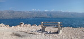 A bench on a rocky shore with a view of mountains and a body of water. - Apartment Tina (Ražanac)