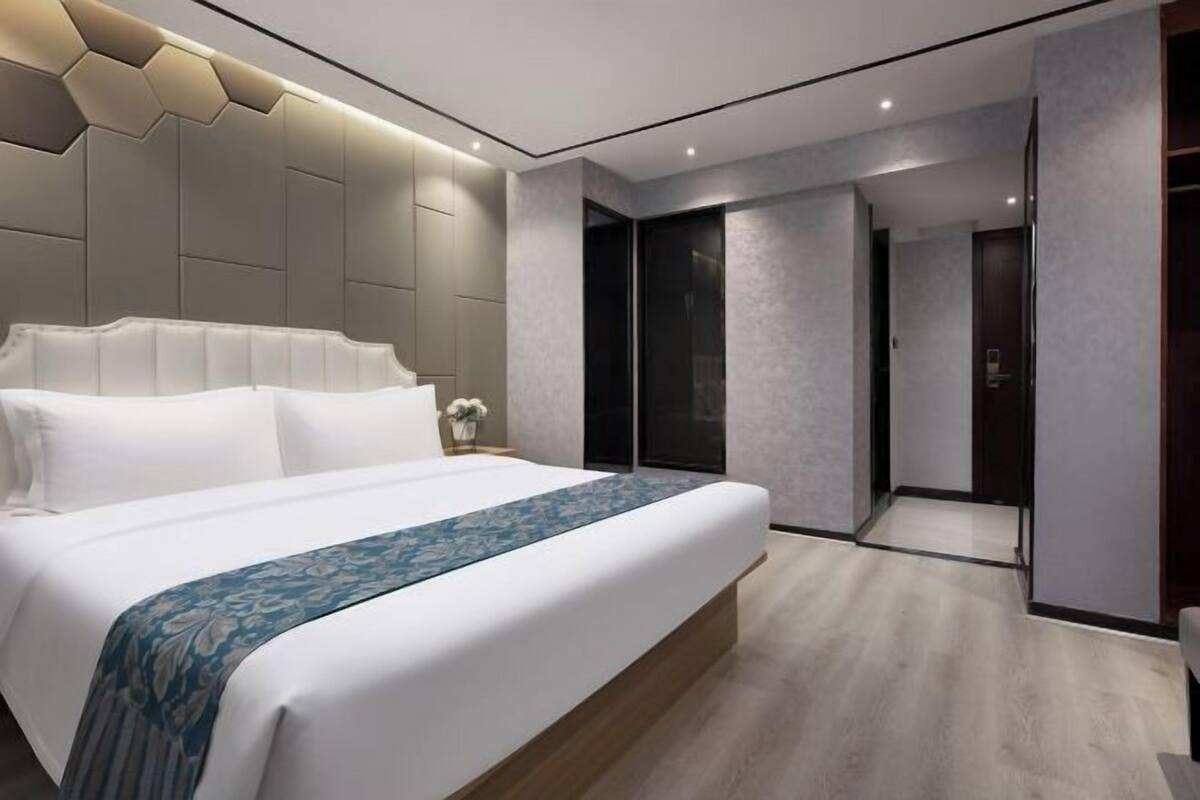 A modern hotel room with a large bed, a headboard with geometric patterns, a walk-in closet, and a bathroom with a glass door.