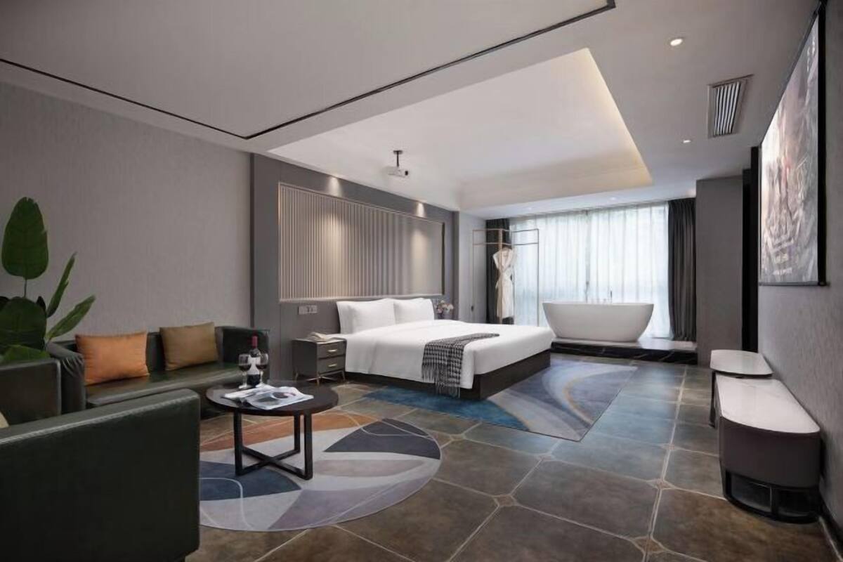 A modern hotel room with a large bed, a sitting area with a sofa and coffee table, a bathtub, and a large window with curtains.