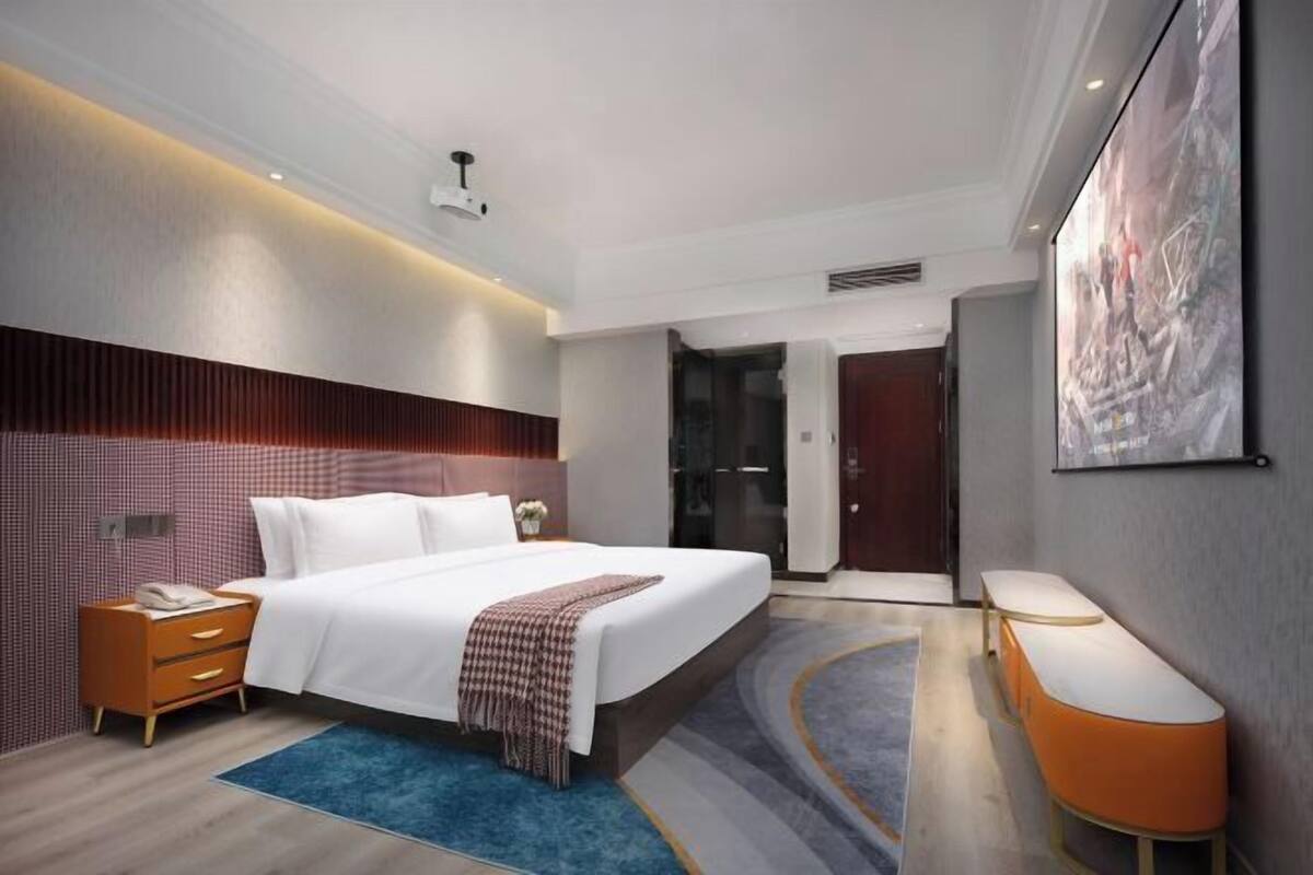 A modern hotel room with a large bed, a bedside table, a desk, and a large window.