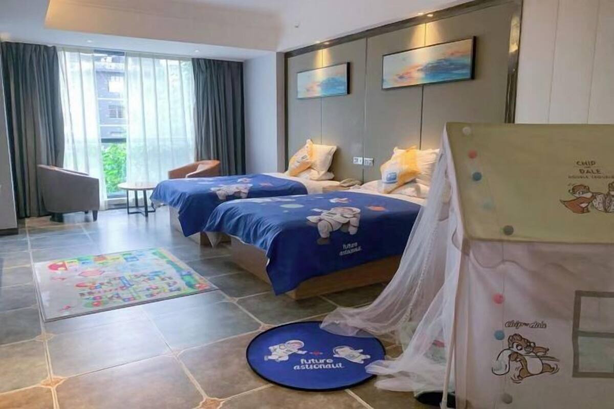 A hotel room with two beds, a small table, a chair, a large window with curtains, and a view of greenery outside.