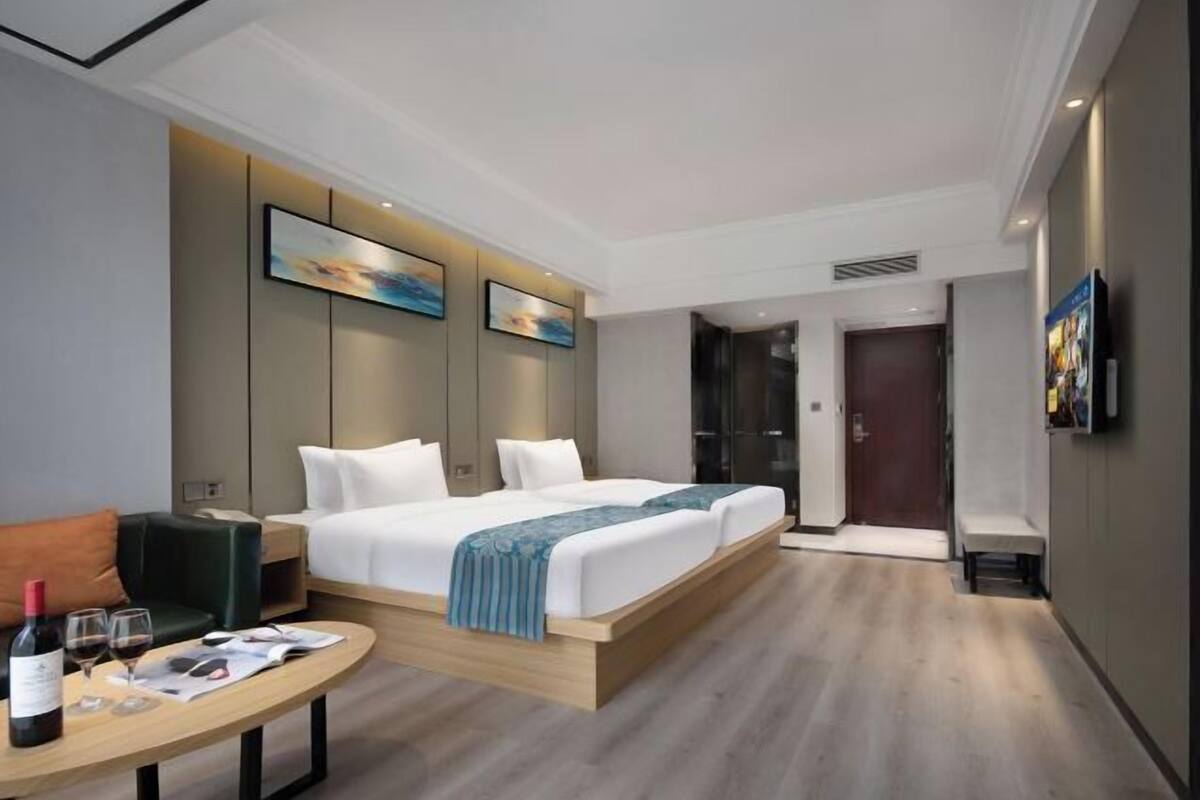 A modern hotel room with a large bed, a small table with a bottle and glasses, a sofa, and artwork on the walls.