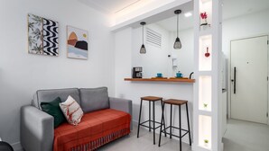 A modern living room with a grey sofa, a red blanket, and two bar stools. There is a kitchen area with a coffee machine and a shelf with decorative items. - 10 Minutes From Flamengo Beach Next to the Subway Ct404 (Rio de Janeiro)
