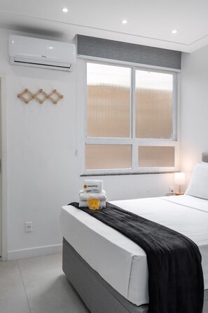 Ein modernes Hotelzimmer mit einem großen Bett, einem Fenster mit Jalousien, einer Klimaanlage und einem kleinen Tisch mit Lampe und einer Ananas-Packung.