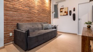 A modern living room with a brick wall, a grey sofa, a small table with a lamp, and a door with a plant on the right. - 7 Minutes From Copacabana Beach Stc504 (Rio de Janeiro)