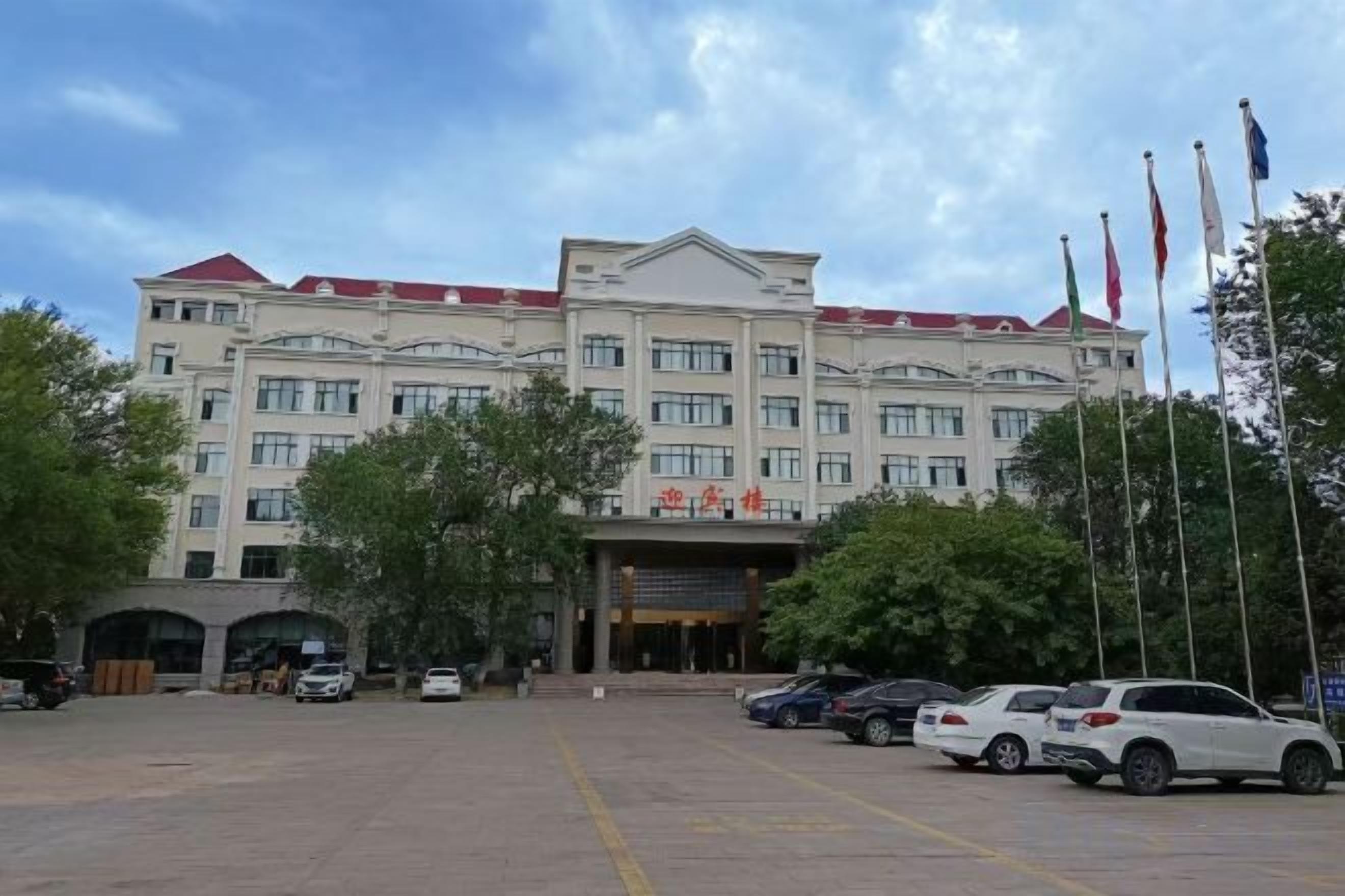 Un hôtel avec une entrée principale, plusieurs drapeaux et un parking où sont garées plusieurs voitures.