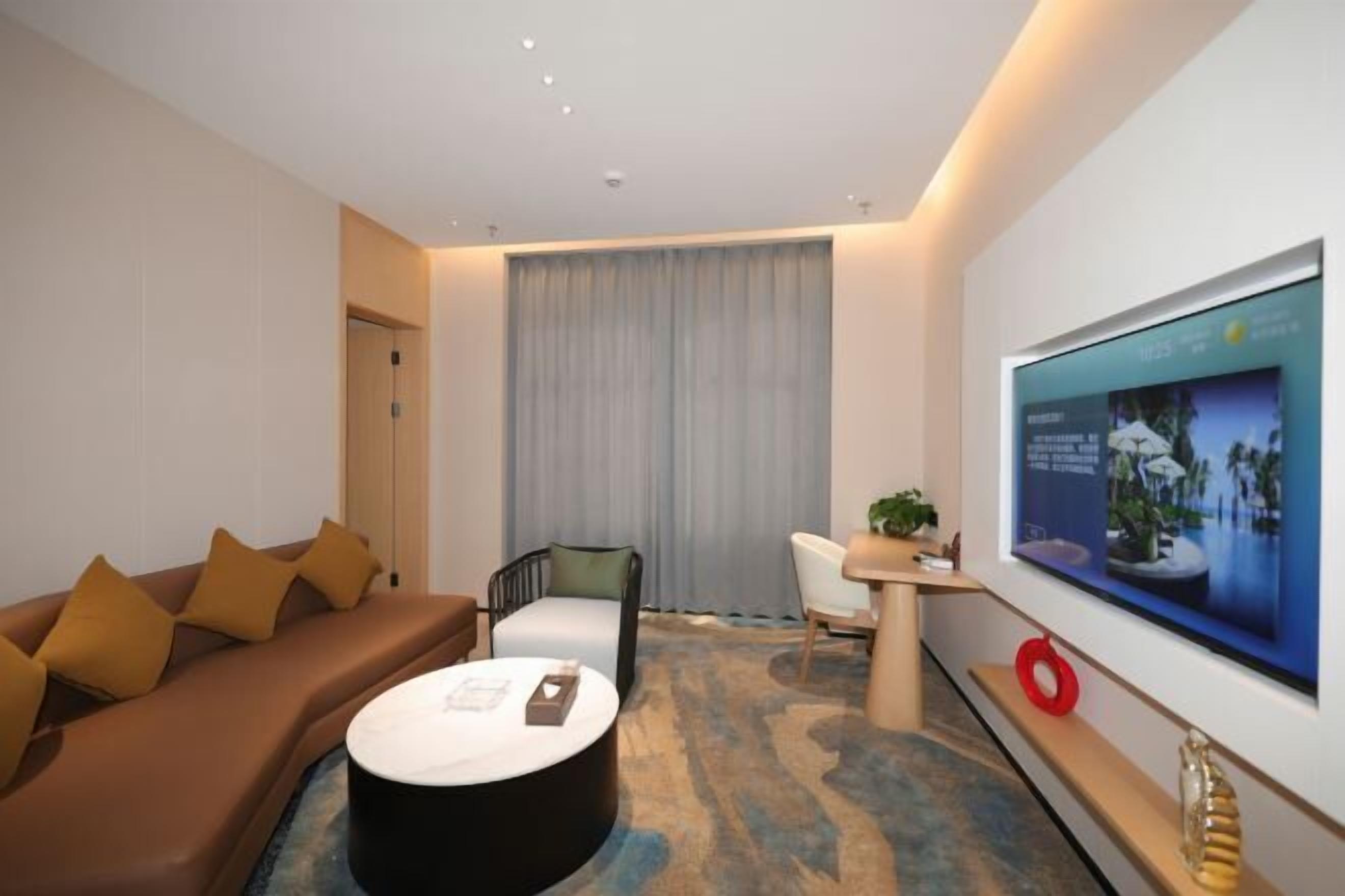 A modern hotel room with a sofa, a round coffee table, a TV mounted on the wall, and a large window with curtains.