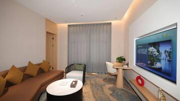 A modern hotel room with a sofa, a round coffee table, a TV mounted on the wall, and a large window with curtains.