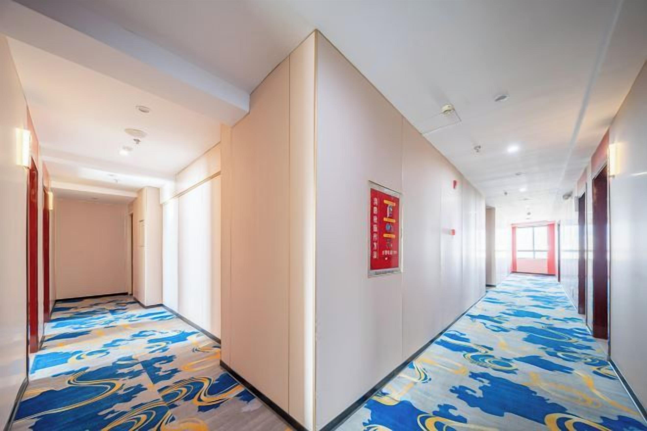 A hotel corridor with a patterned carpet, a red exit sign, and a door at the end.