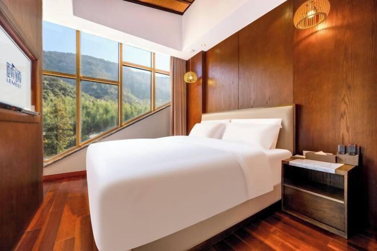 Una habitación de hotel moderna con una cama grande, paneles de madera, una mesita de noche y vistas a un bosque a través de amplias ventanas.