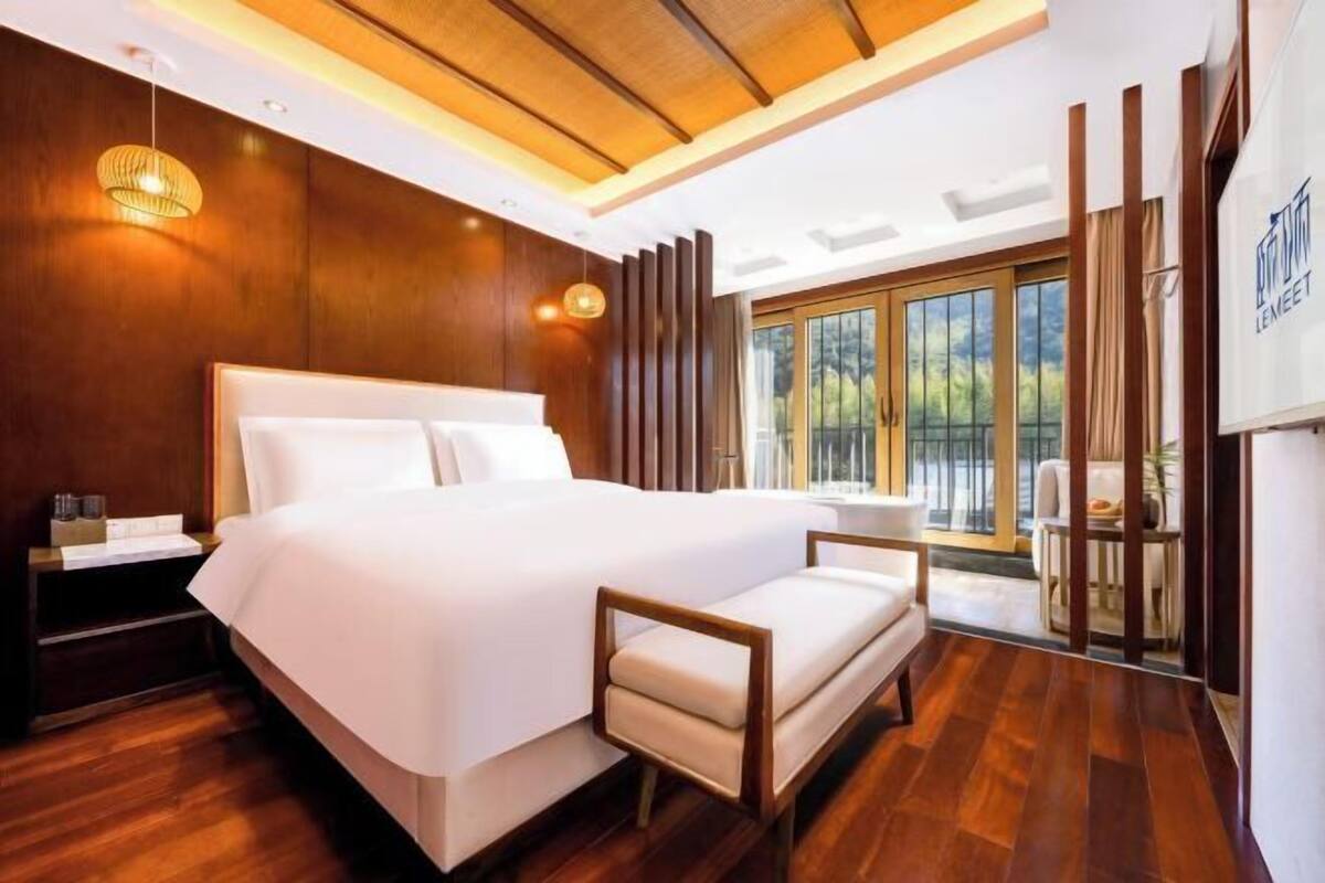 Habitación de hotel moderna con una cama grande, suelo de madera, un pequeño sofá, una mesita de noche y un ventanal con vistas a zonas verdes.