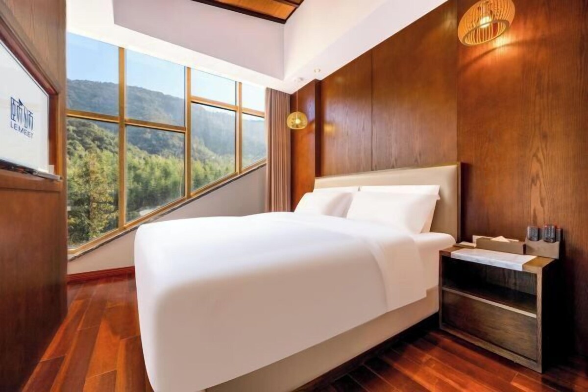 Une chambre d’hôtel moderne dotée d’un grand lit, de lambris en bois, d’une table de chevet et offrant une vue sur une forêt grâce à de grandes fenêtres.