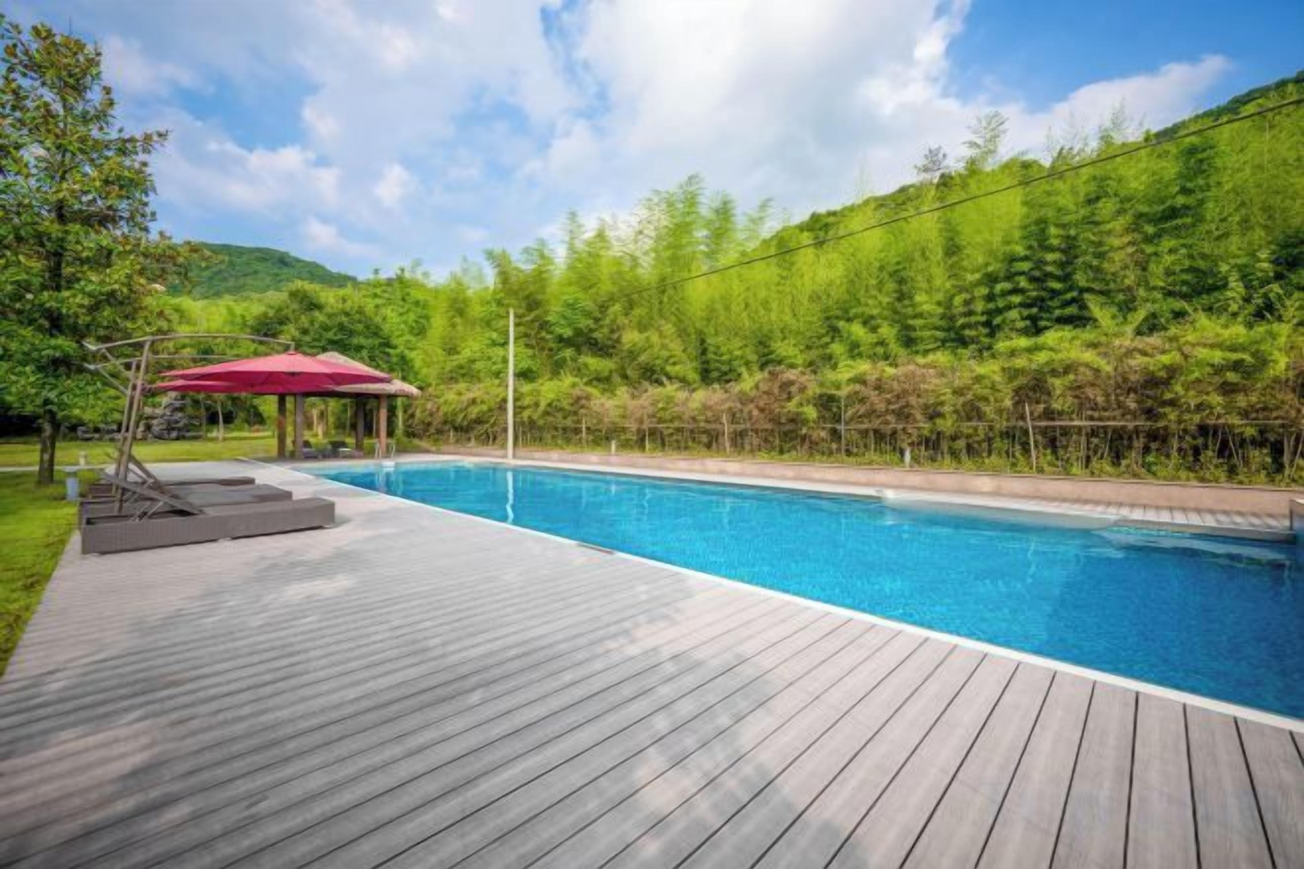 A swimming pool with a wooden deck, surrounded by greenery and a red umbrella.
