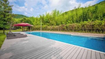 A swimming pool with a wooden deck, surrounded by greenery and a red umbrella.
