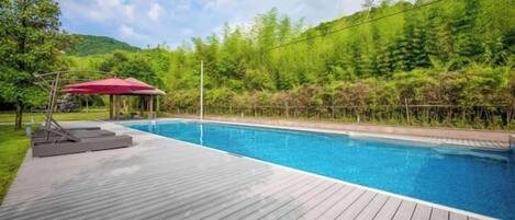 A swimming pool with a wooden deck, surrounded by greenery and a red umbrella.