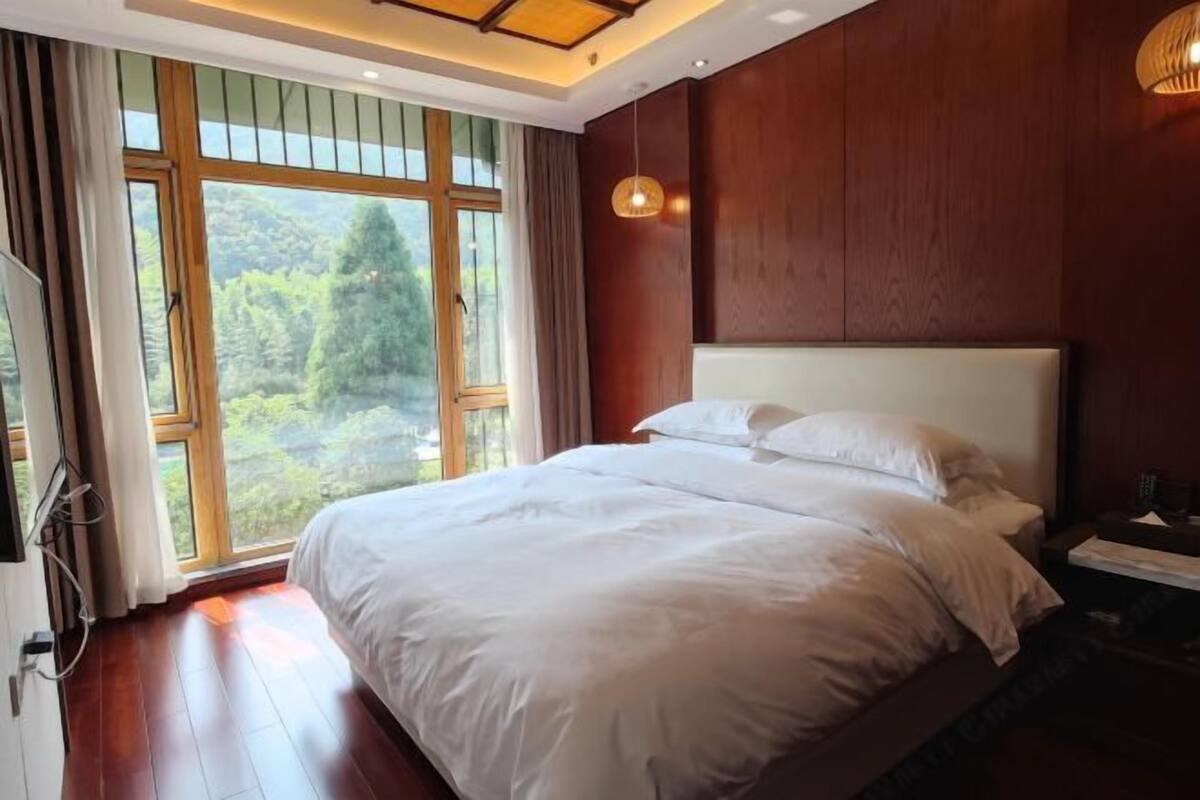 Un dormitorio con una cama grande, paredes de madera, un ventanal con vistas a la vegetación y una mesita de noche con lámpara.