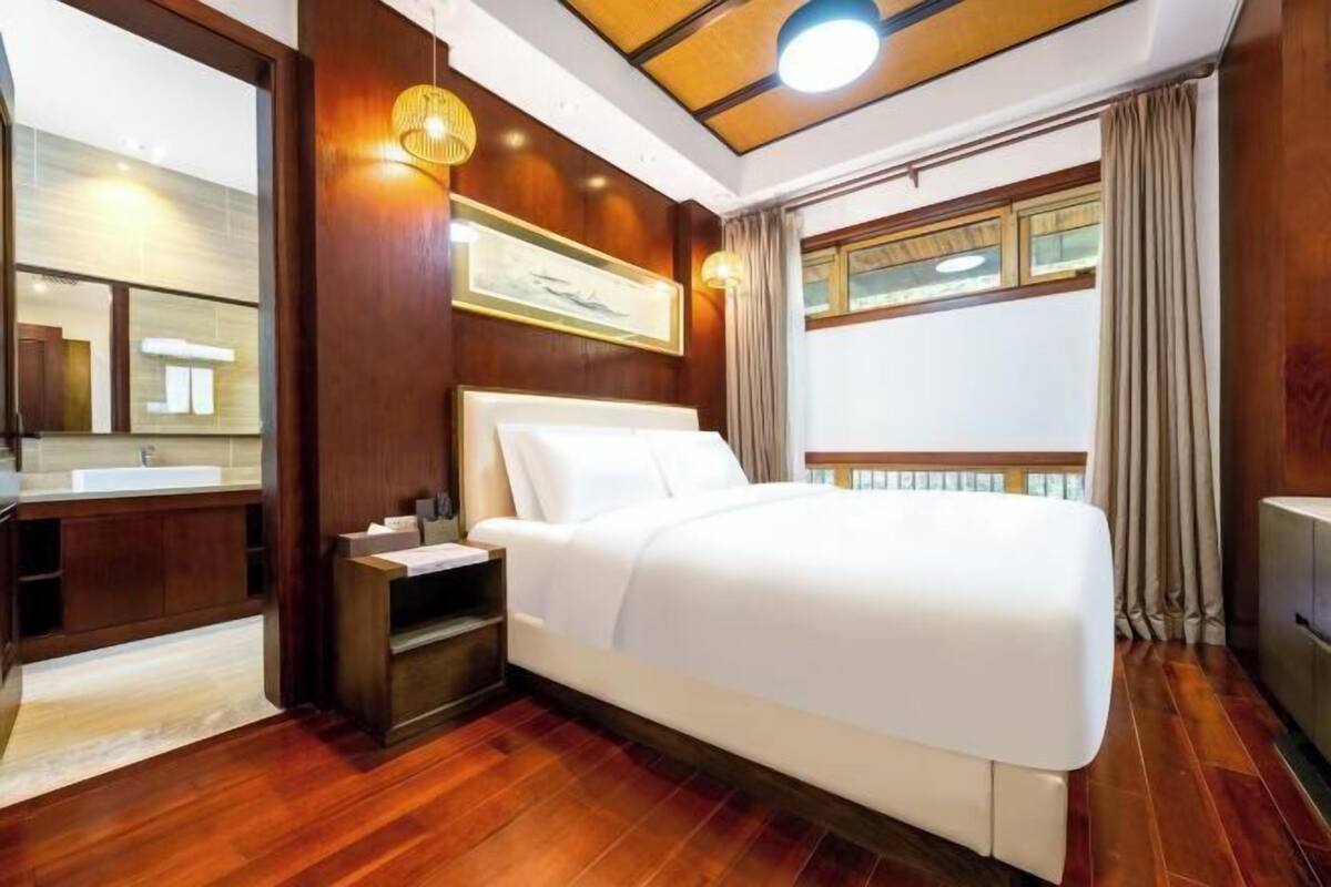 Habitación de hotel con una cama grande, piso de madera, baño con lavamanos y espejo, y ventana con cortinas.