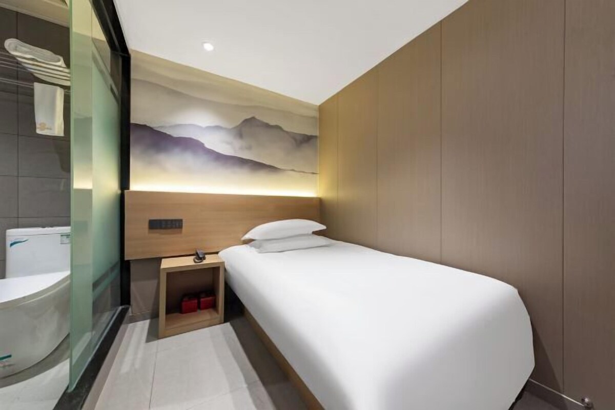 A modern hotel room with a bed, a nightstand, a wall-mounted light, a bathroom with a toilet, and a glass partition.