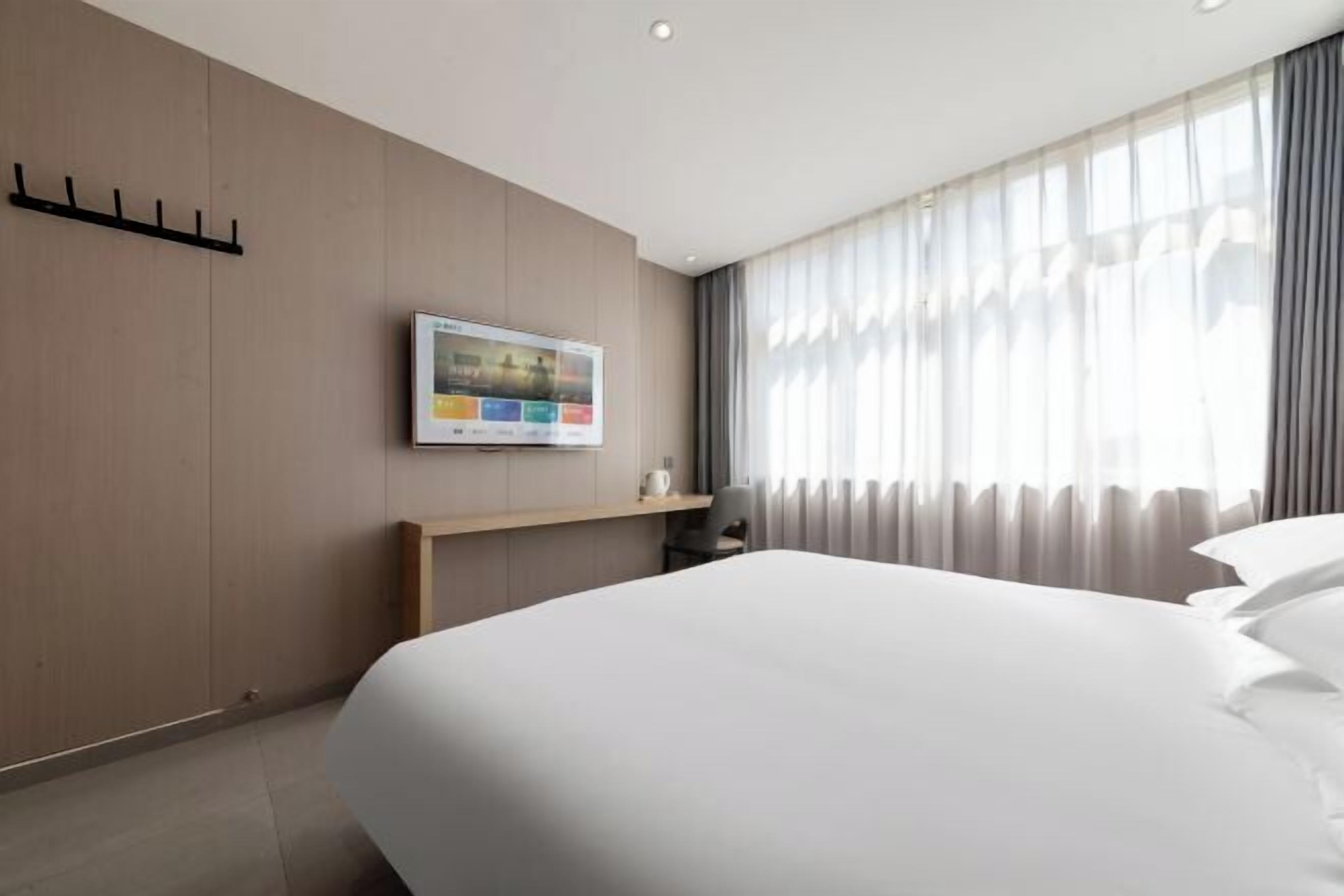 Una habitación de hotel con una cama grande, un televisor de pantalla plana en la pared, una mesita de noche de madera y una ventana con cortinas translúcidas.