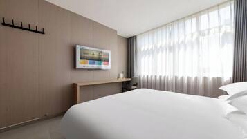 A hotel room with a large bed, a flat-screen TV mounted on the wall, a wooden nightstand, and a window with sheer curtains.