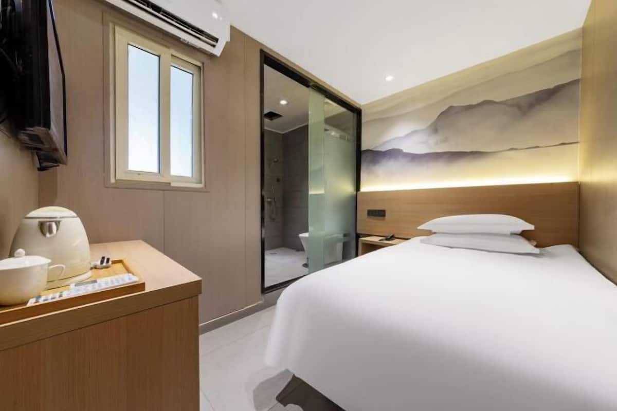 Una habitación de hotel moderna con una cama, un escritorio con una tetera, un espejo y un baño visible a través de una puerta de vidrio.