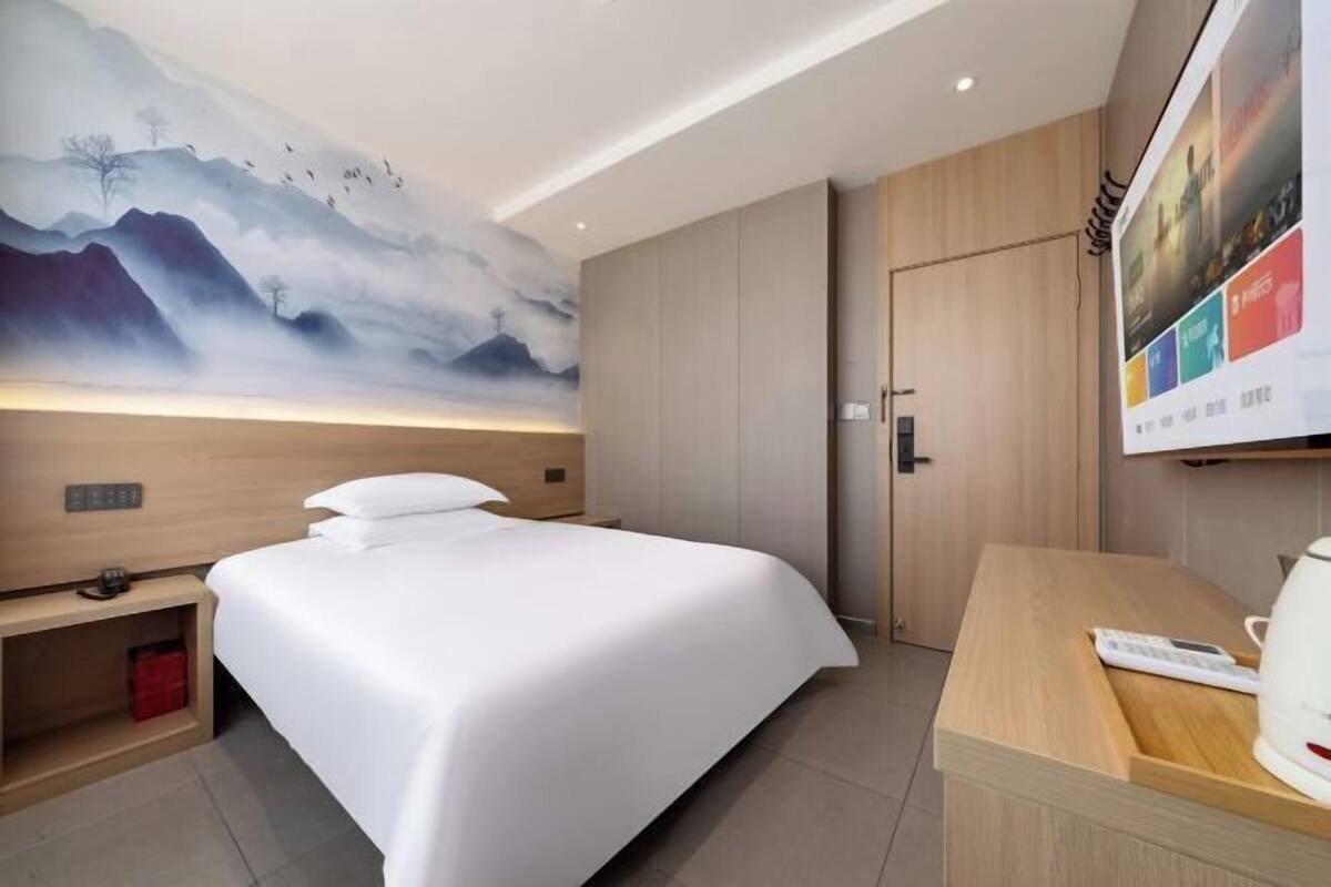Habitación de hotel con una cama grande, cabecera de madera, mesita de noche, escritorio con hervidor y televisor montado en la pared.