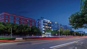 A multi-story building with a red facade and a blue-lit upper section, surrounded by greenery and a well-maintained road. - Sangpin E-sports Hotel (Wenzhou Guoxi Branch) (Wenzhou)