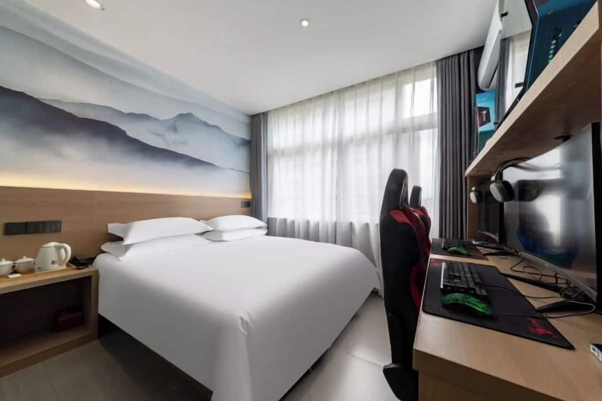 Una habitación de hotel con una cama grande, un escritorio con computadora y auriculares, un televisor y un mural en la pared.