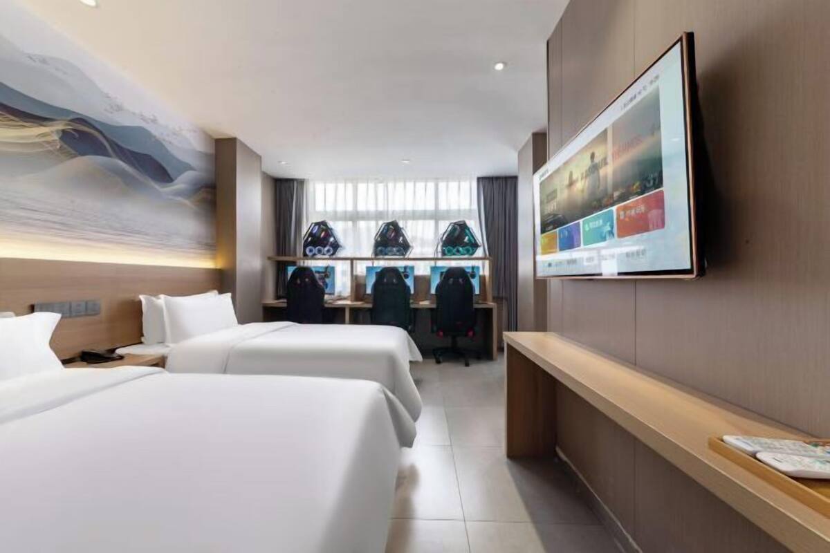 Una habitación de hotel con dos camas, un escritorio con computadoras y un televisor grande en la pared.