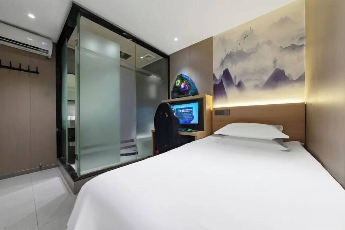 Una habitación de hotel moderna con una cama, un escritorio con una computadora, una puerta de cristal que da a un balcón y una pared con un cuadro de montañas.