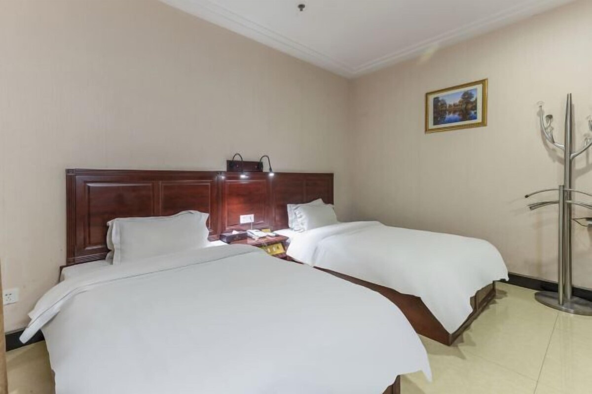 A hotel room with two beds, a wooden headboard, a nightstand, a lamp, a coat rack, and a framed picture on the wall.