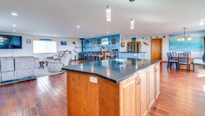 A spacious living area with a wooden floor, a large island with a black countertop, and a dining area with chairs and a table. - Ocean Shores Retreat w/ Fire Pit - Walk to Beach! (Ocean Shores)