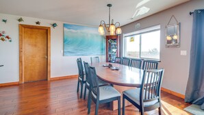 A dining area with a wooden table and chairs, a painting of a wave on the wall, and a window with a view. - Ocean Shores Retreat w/ Fire Pit - Walk to Beach! (Ocean Shores)