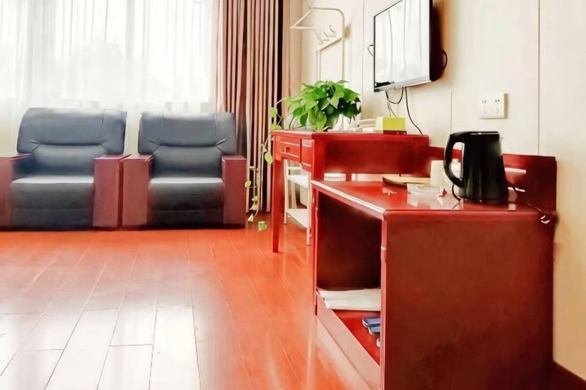A hotel room with a wooden floor, a red desk, a black coffee maker, a television on the wall, and two grey chairs.