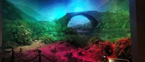 Una exhibición de museo con un puente de piedra, abundante follaje y una zona alfombrada de color rojo.