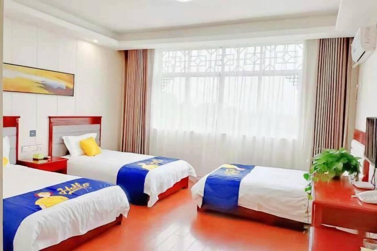 A hotel room with two beds, a desk, a chair, a plant, and a window with curtains.