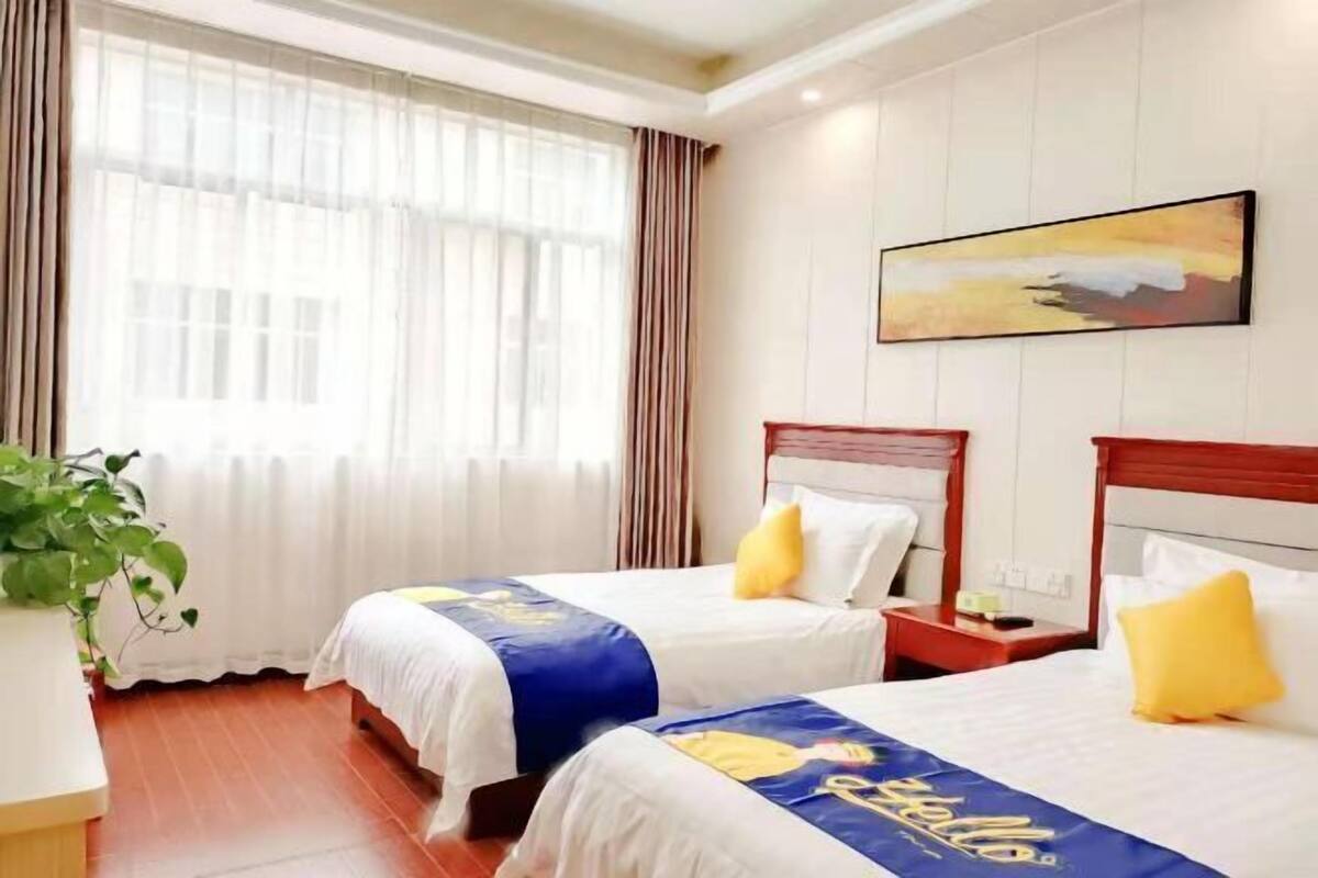 A hotel room with two beds, a large window with curtains, a painting on the wall, and a plant.