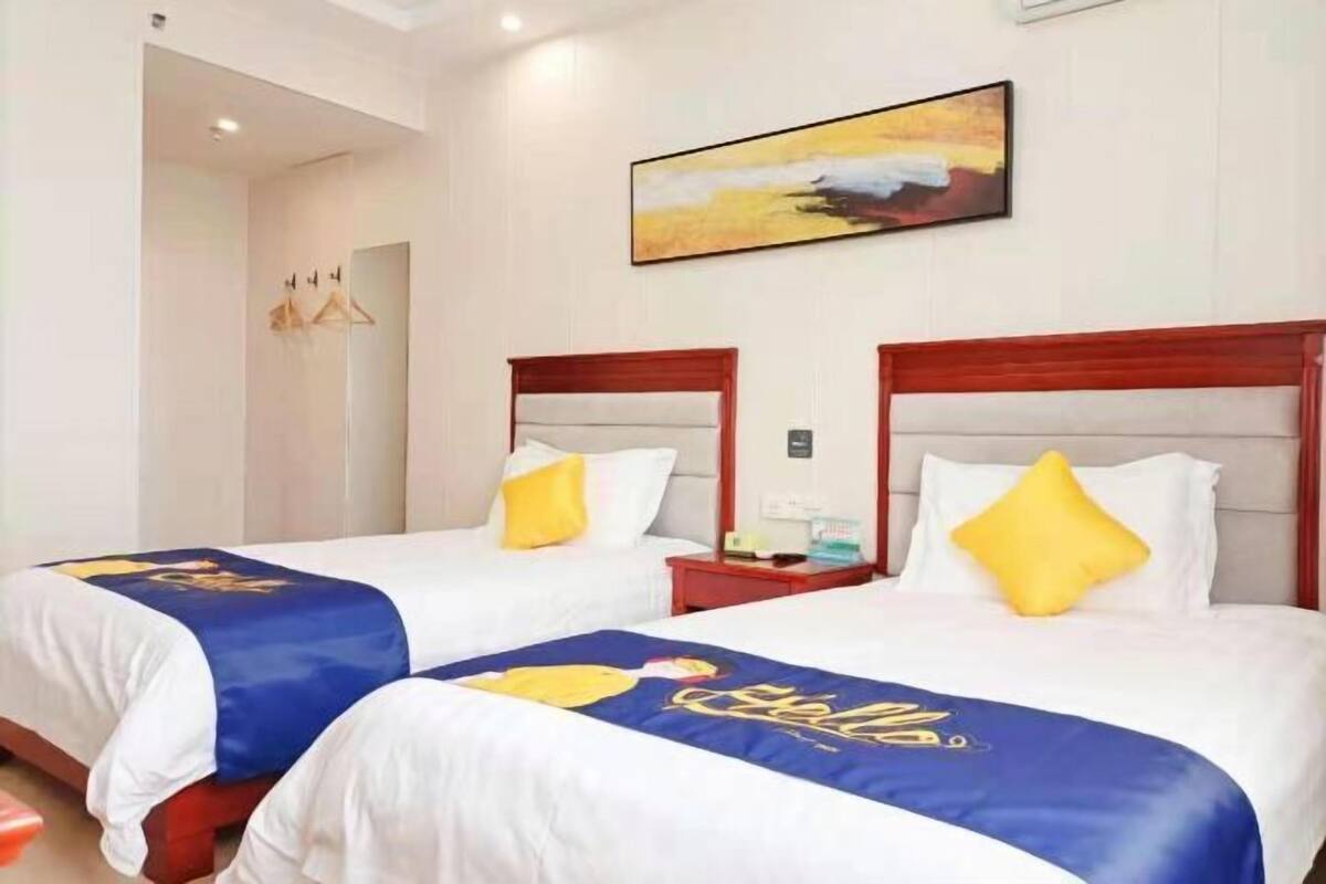 A hotel room with two beds, each with a blue and white bedspread and a yellow pillow. There is a painting on the wall above the beds and a red nightstand between them.
