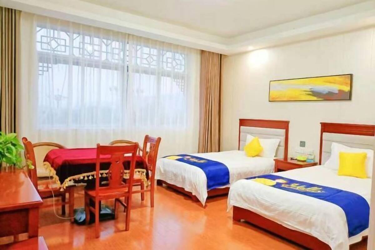 A hotel room with two beds, a dining table with chairs, a potted plant, and a painting on the wall.