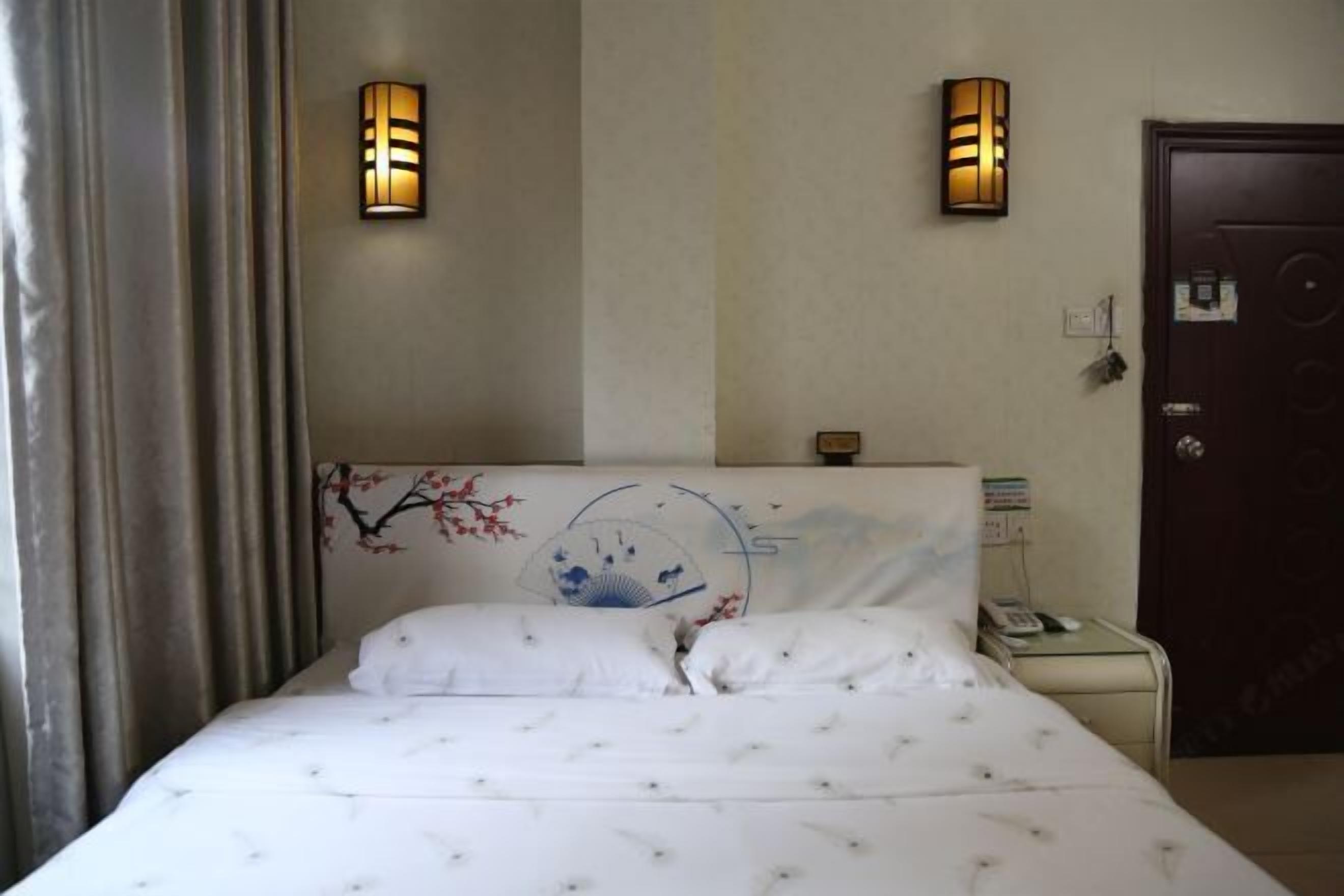 Quarto de hotel com cama, duas luminárias na parede, mesa de cabeceira com telefone e porta com tranca.