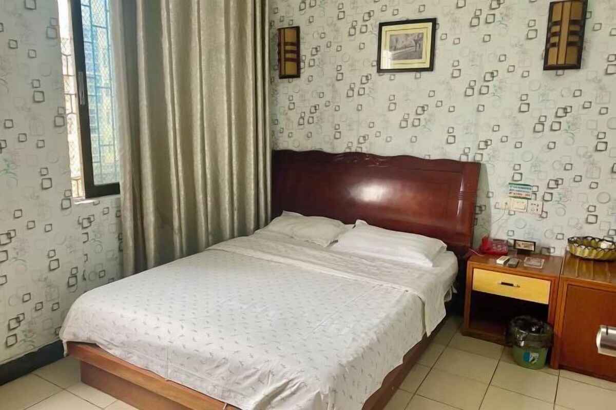 A bedroom with a bed, curtains, a wooden headboard, a nightstand, a trash bin, and patterned wallpaper.