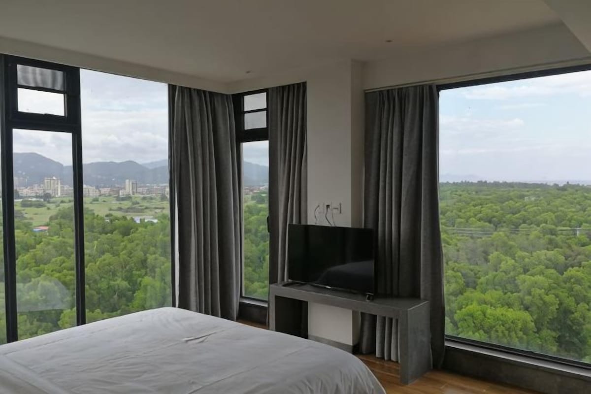 A bedroom with a large bed, a flat-screen TV, and a city view through large windows.