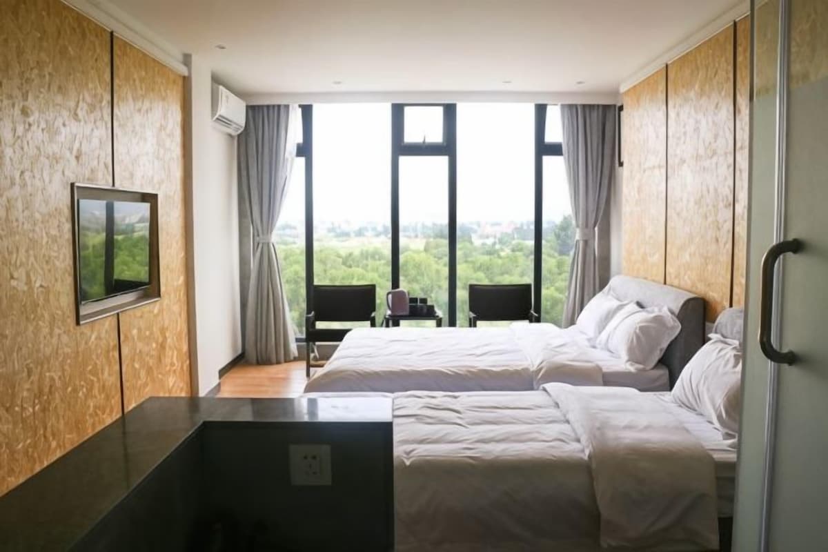 A hotel room with two beds, a TV, and a view of greenery.