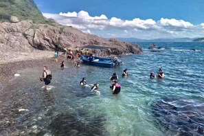 A rocky shoreline with clear water, a small boat, and people swimming and wading. - Destiny (Huidong)