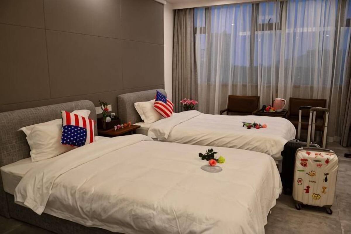 A hotel room with two beds, a window with curtains, a small table with a vase of flowers, and a suitcase.