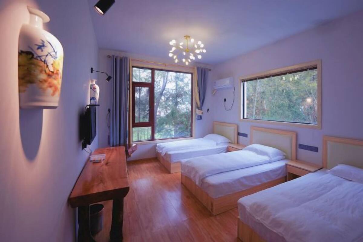 Une chambre avec deux lits, une table en bois, un luminaire fixé au mur et une fenêtre donnant sur des arbres.