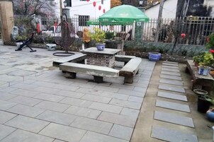 Un espacio al aire libre empedrado con un banco de hormigón, plantas en macetas y un paraguas verde.