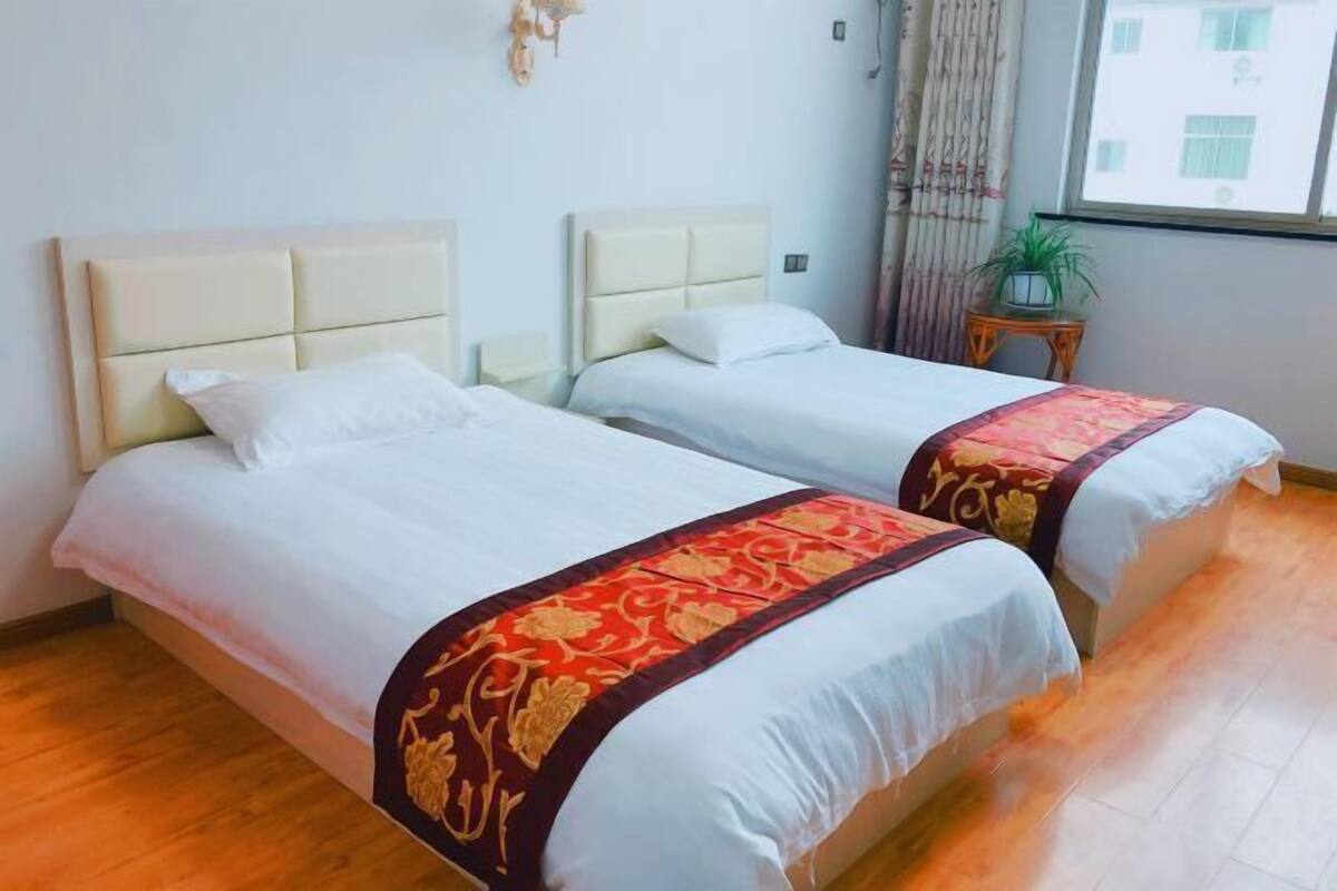 Une chambre d’hôtel avec deux lits, une fenêtre avec des rideaux, une petite table avec une plante et un élément de décoration fixé au mur.