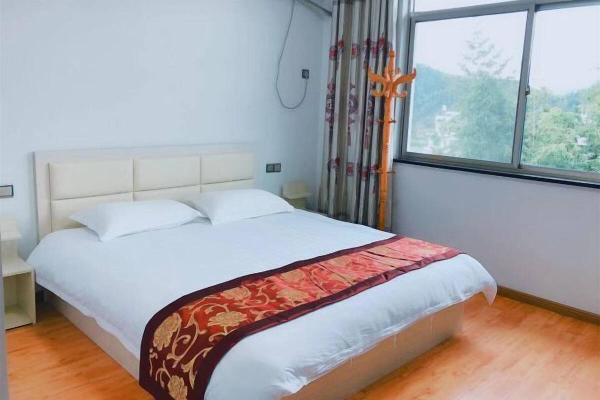 Un lit bien fait, avec une tête de lit blanche, un couvre-lit décoratif rouge et or, et une fenêtre donnant sur un paysage verdoyant.