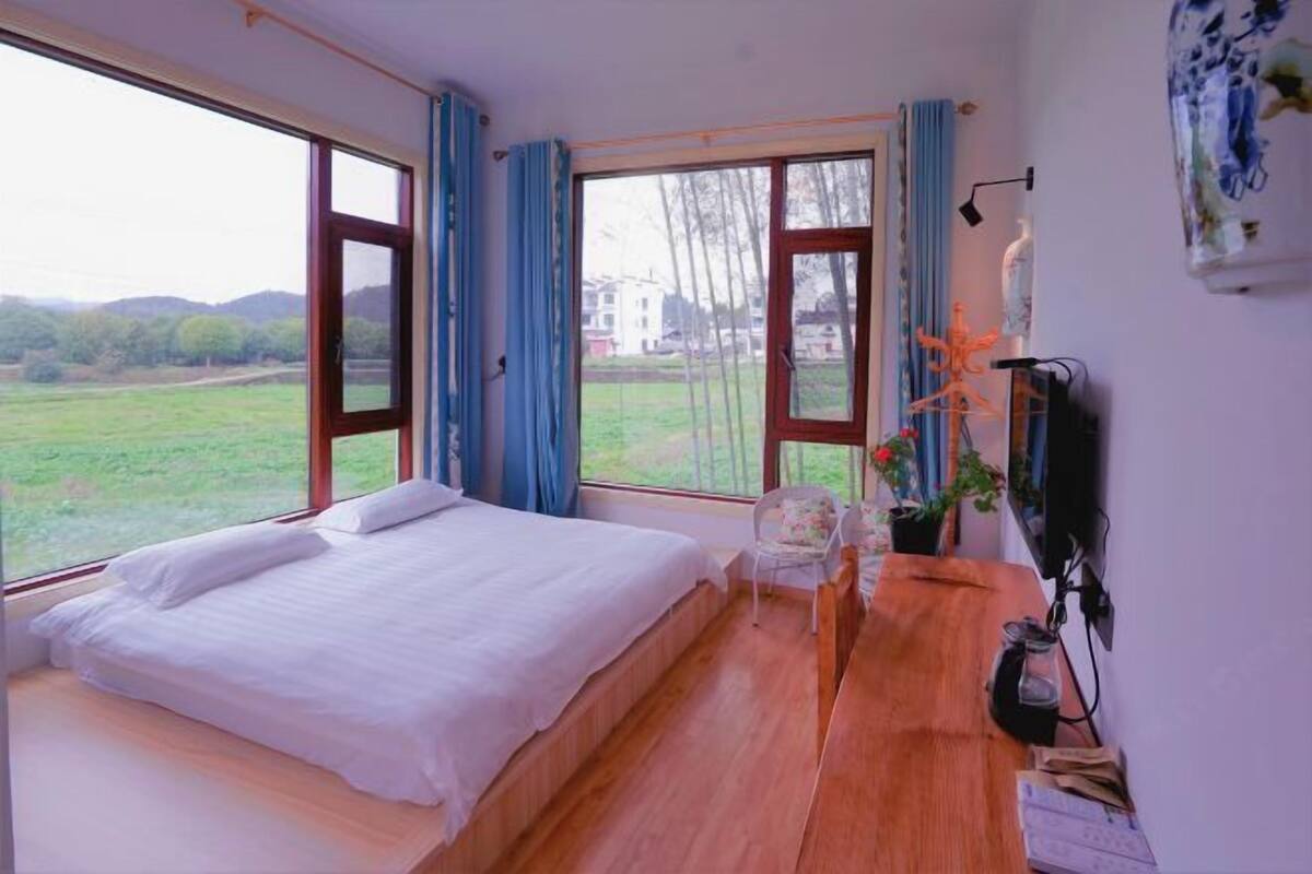Une chambre à coucher comprenant un lit, un bureau, une chaise, un téléviseur et de grandes fenêtres donnant sur un paysage verdoyant.